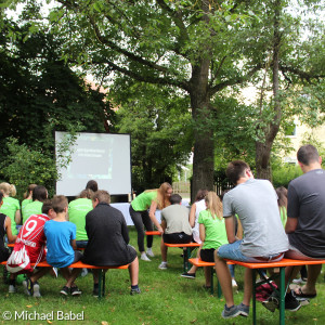 Junge Menschen auf Bierbänken vor einer Leinwand und einem Altar im Garten